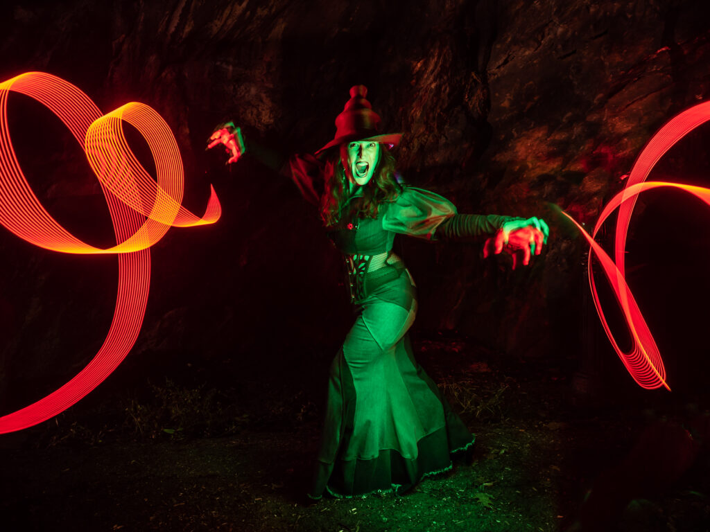 Green lit model in a witch outfit with red light painting trails coming from the hands